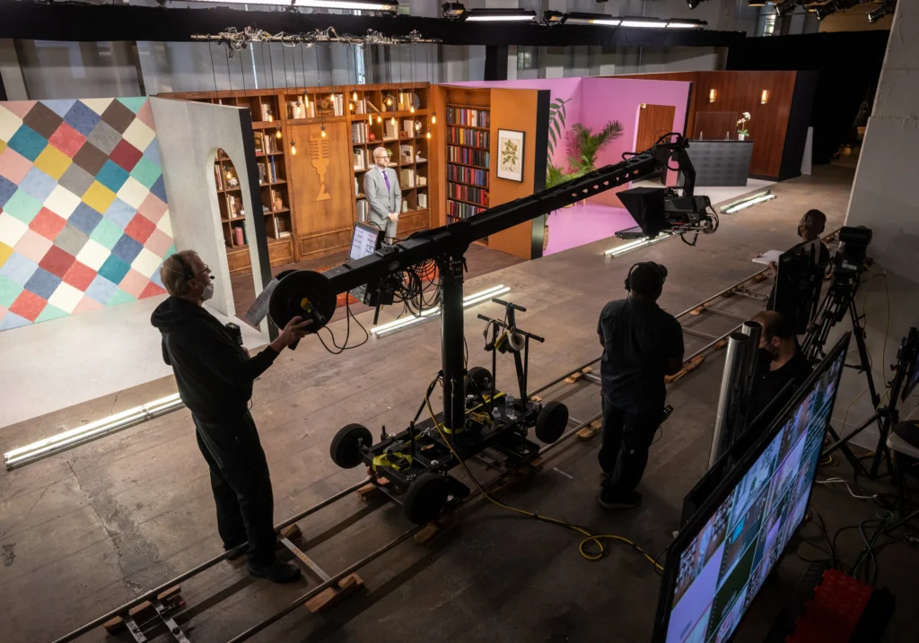 Camera operators stand next to a large camera on tracks as a presenter waits on set.