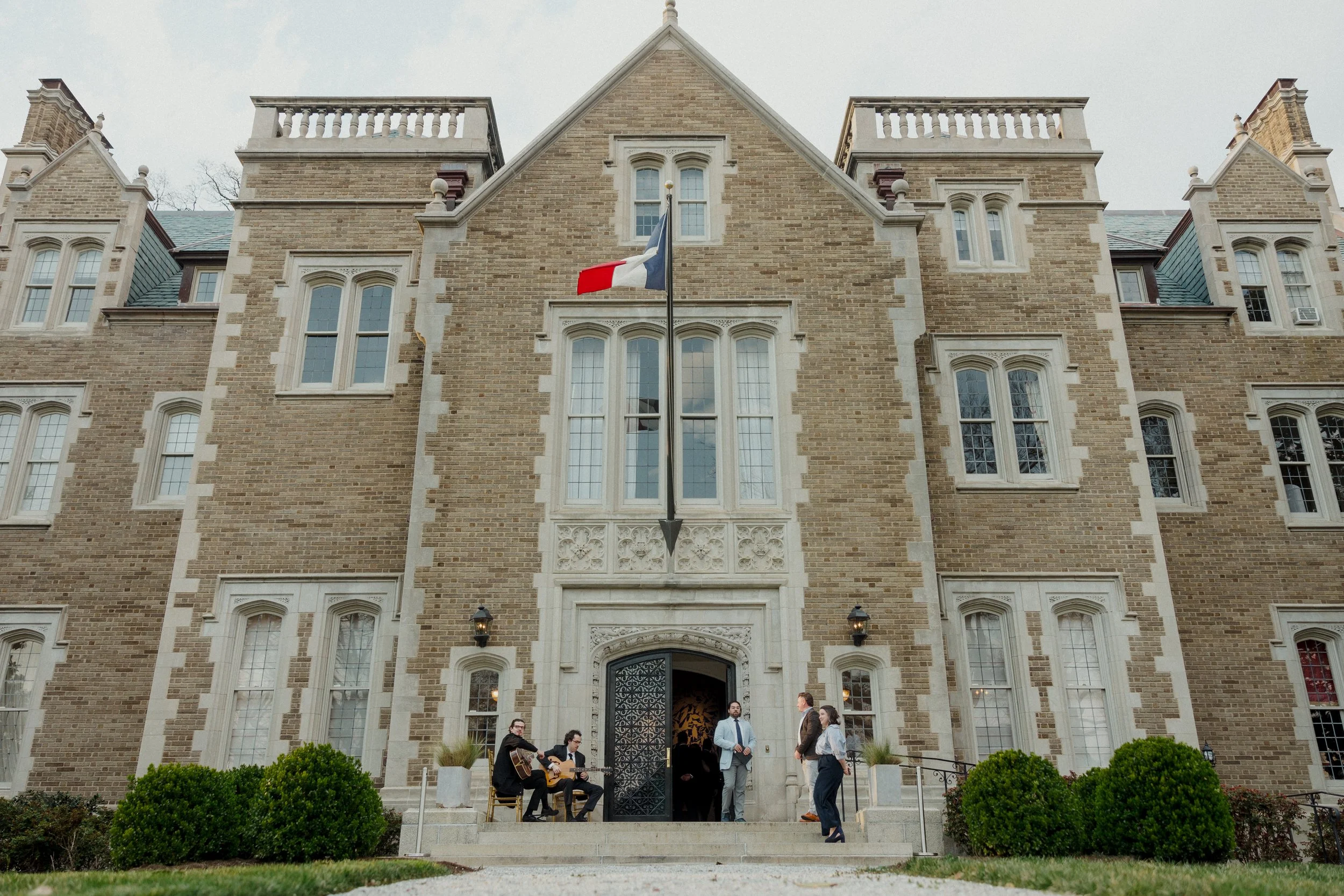Two men play guitars on the front steps of a historic building as guests enter.