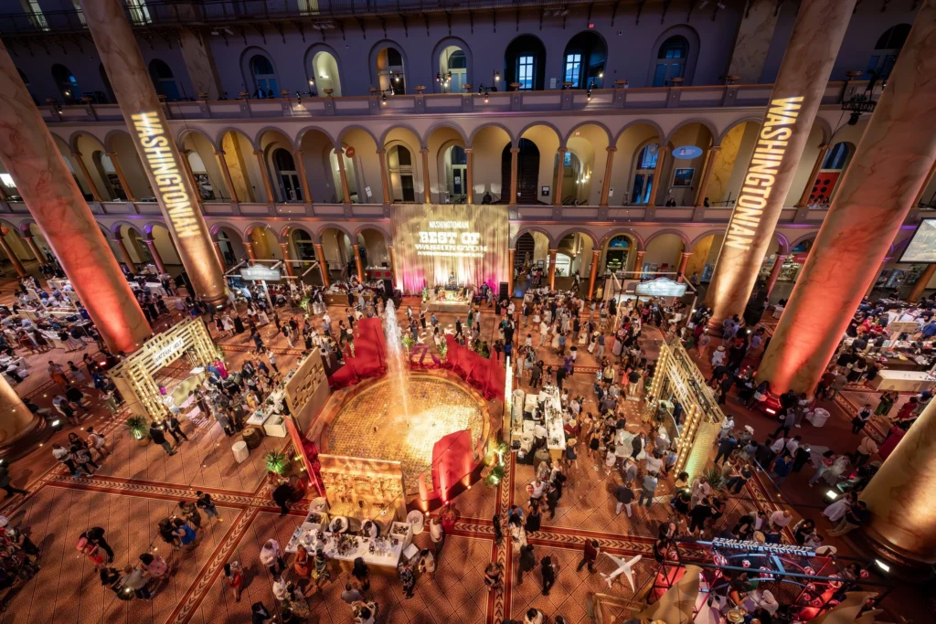 An aerial drone shot of the event space for Best of Washington presented by Washingtonian Magazine. People mingle and wander among stages and huge pillars with red and gold projections.