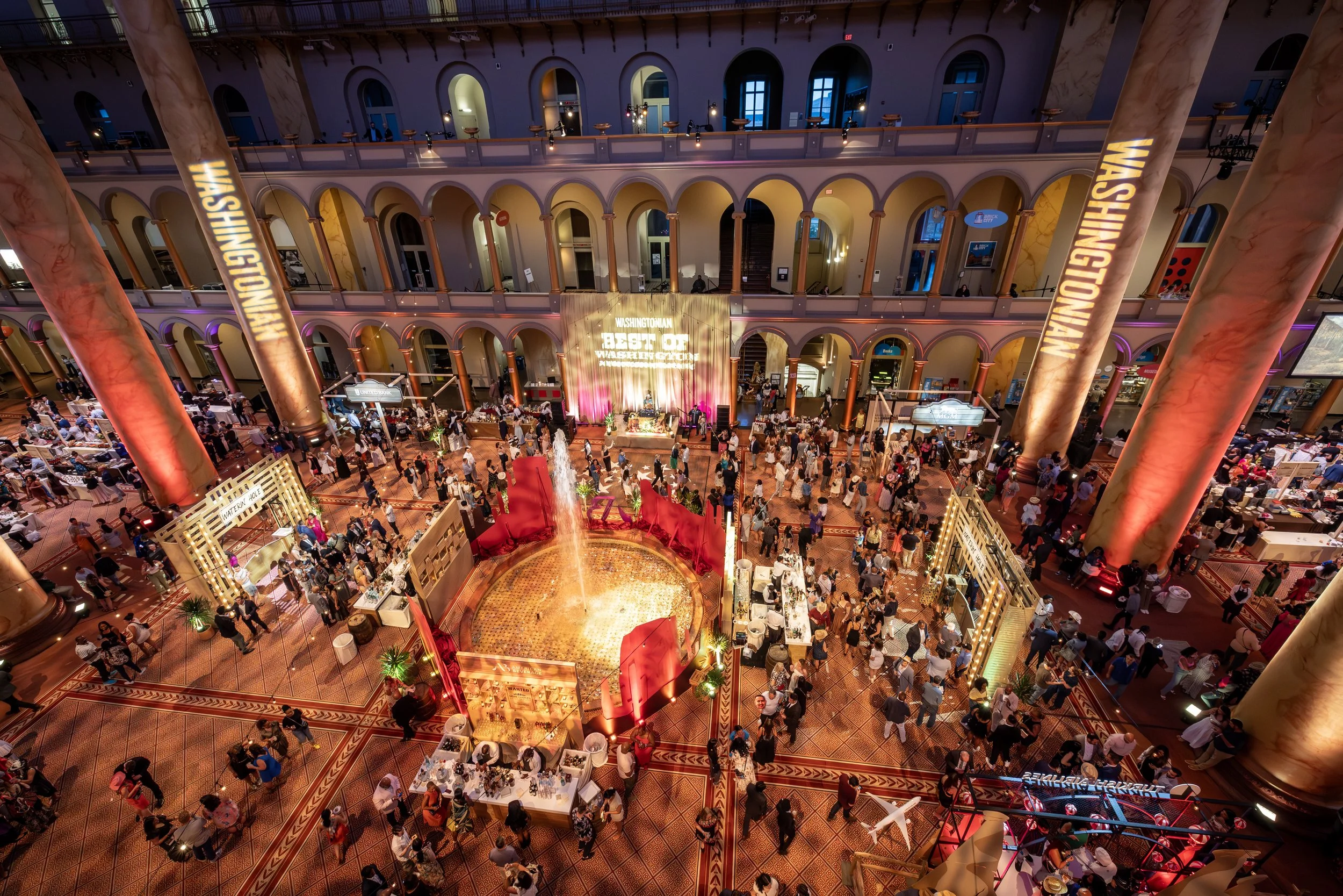 An aerial drone shot of the event space for Best of Washington presented by Washingtonian Magazine. People mingle and wander among stages and huge pillars with red and gold projections.
