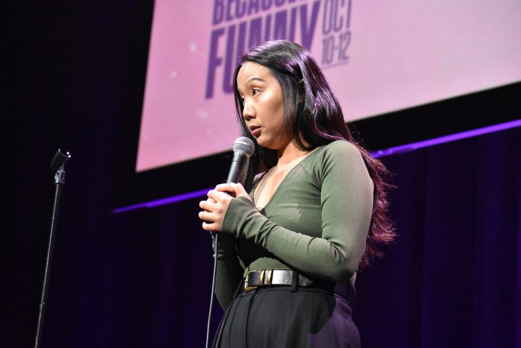 A woman holds a mic onstage at the 2025 "Because They're Funny" Comedy Festival