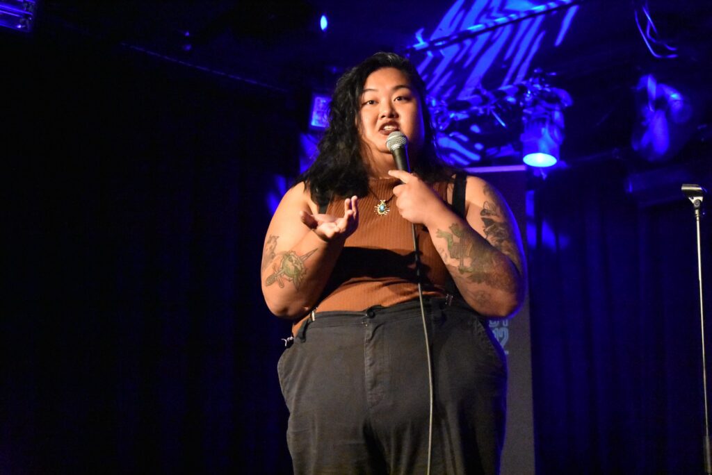 A woman holds a mic onstage at the 2025 "Because They're Funny" Comedy Festival