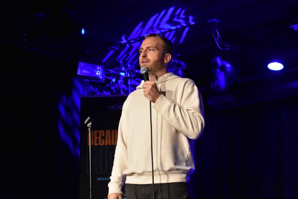 A man holds a mic onstage at the 2025 "Because They're Funny" Comedy Festival