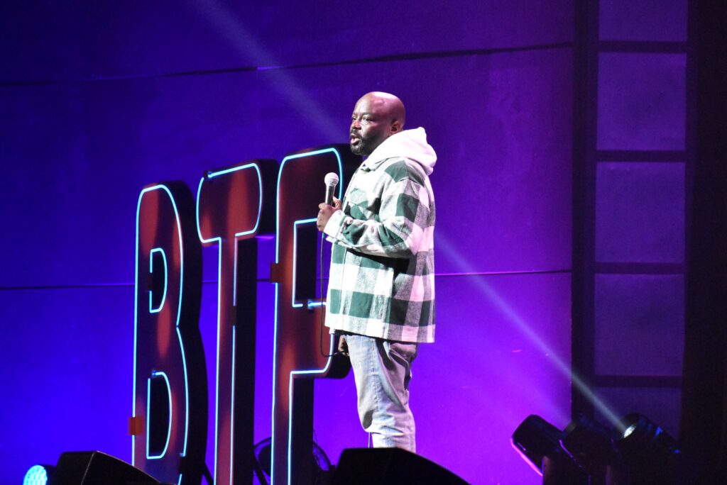 A man holds a mic onstage at the 2025 "Because They're Funny" Comedy Festival
