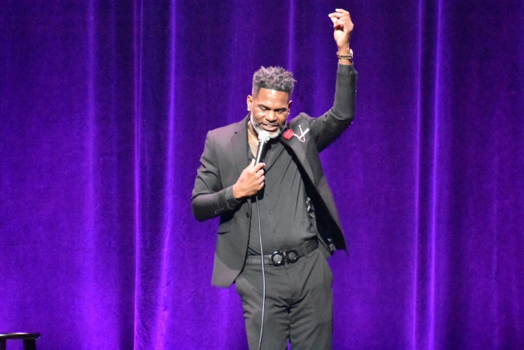A man raises his hand in the air, holding a mic onstage at the 2025 "Because They're Funny" Comedy Festival