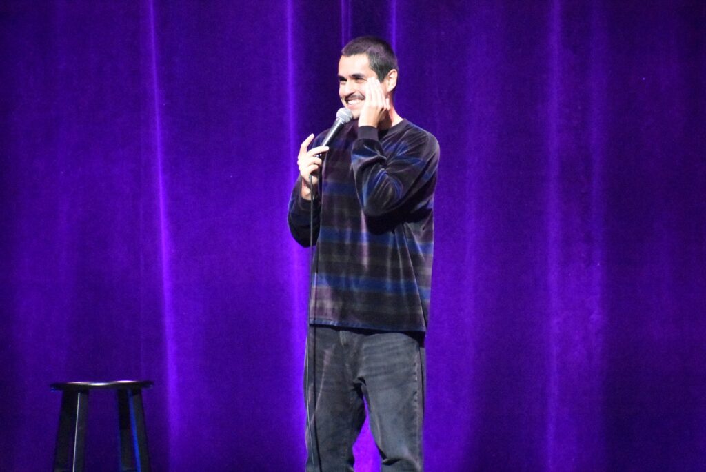 A man smiles onstage in front of a mic at the 2025 "Because They're Funny" Comedy Festival