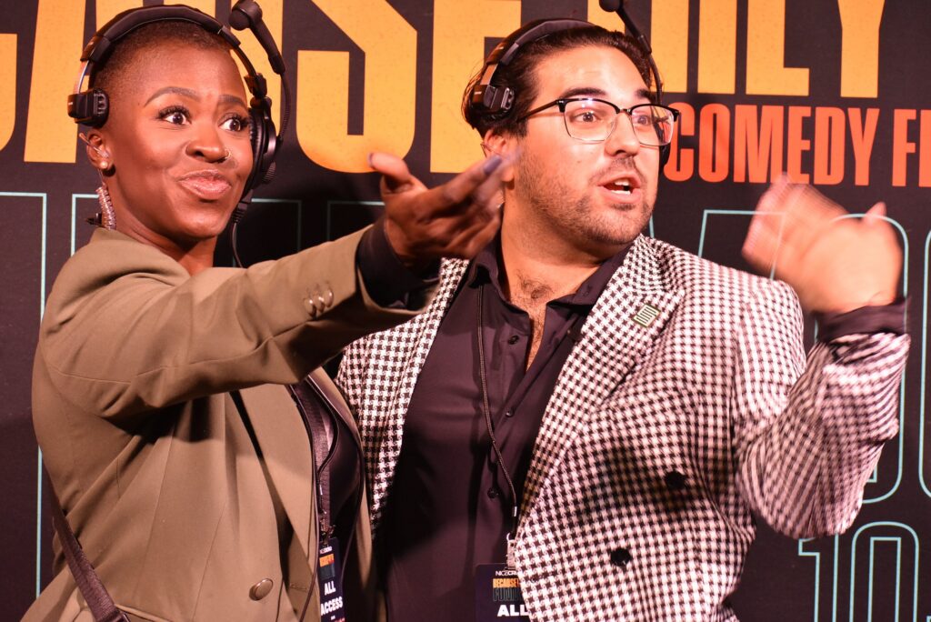Two people beckon someone offscreen to join them in a photo at the 2025 "Because They're Funny" Comedy Festival