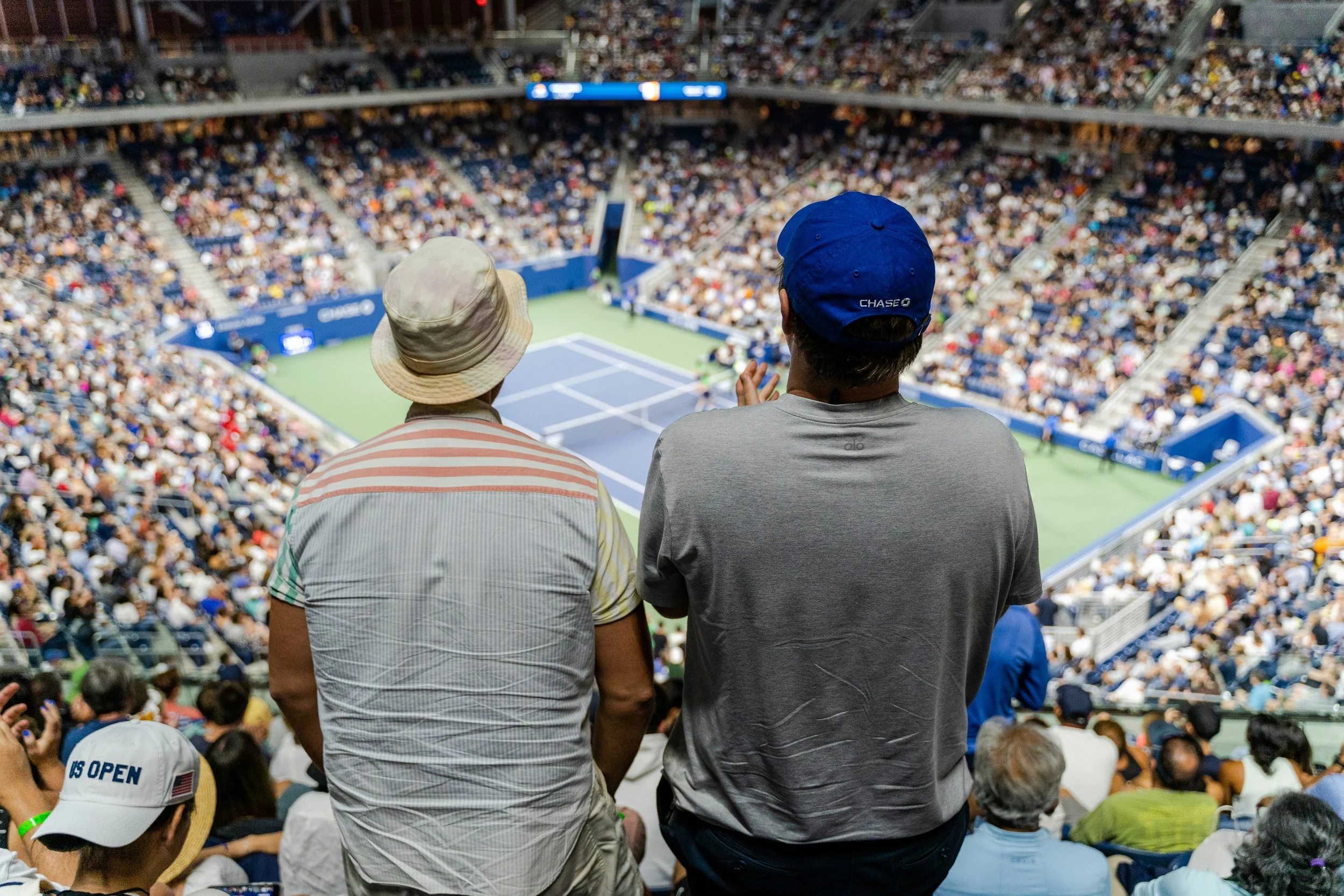 Two men sit and watch a tennis match in a crowded stadium