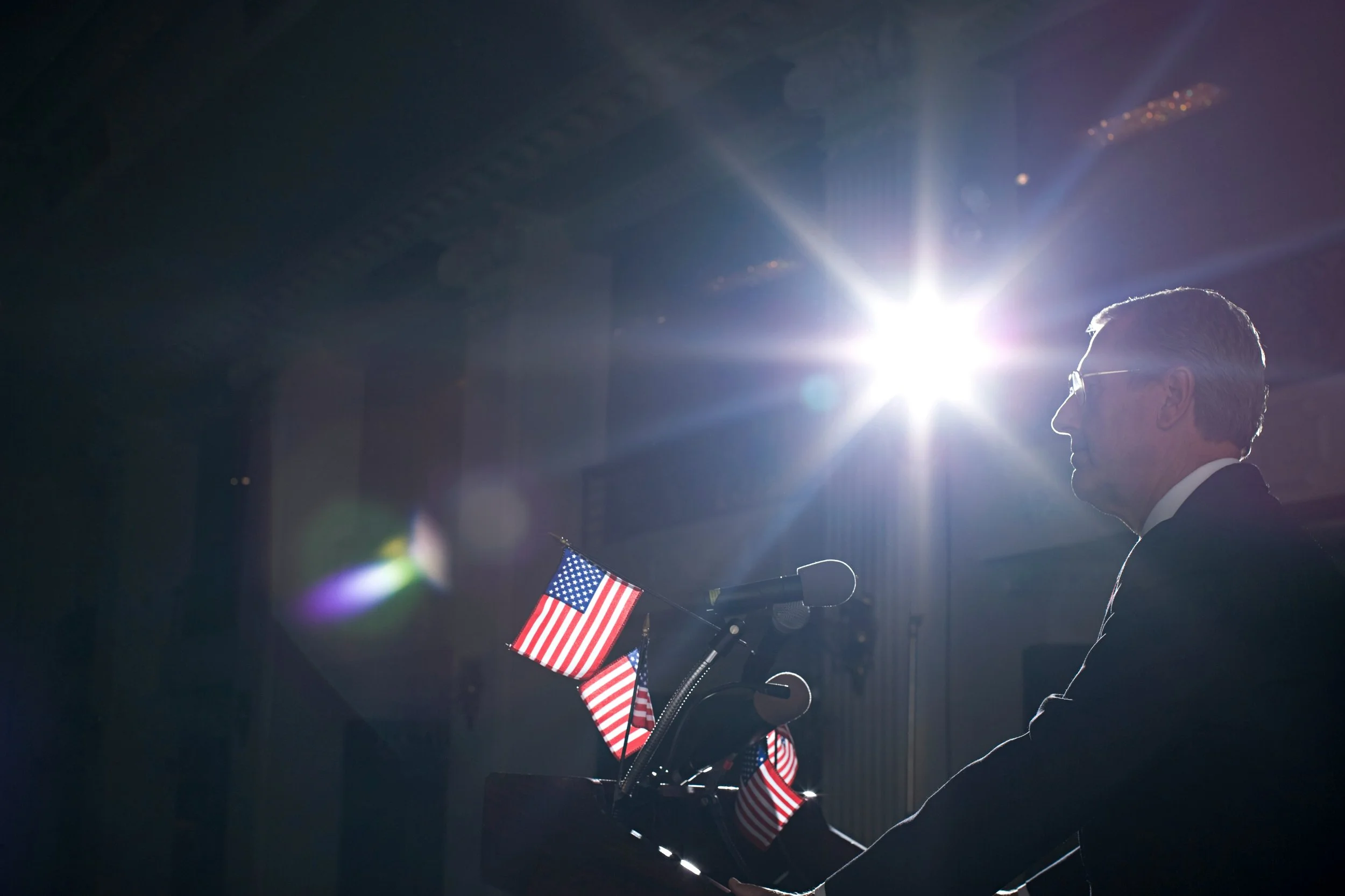 A man silhouetted by lights speaks at a podium covered in small American flags.