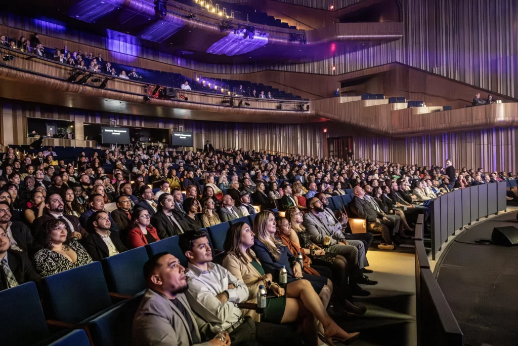 A grand auditorium filled with audience members, all watching the stage.