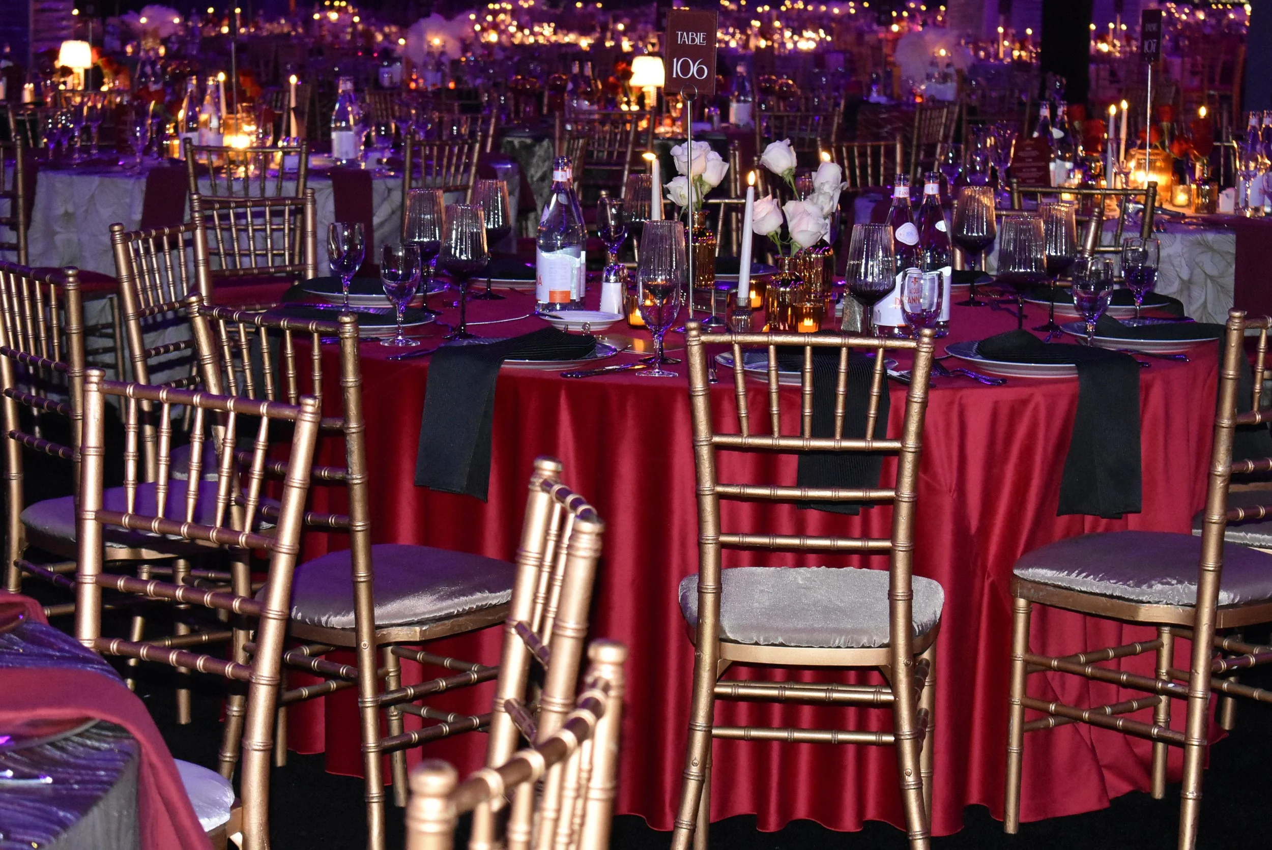 Gold chairs surround tables draped with red tablecloths and set with elegant place settings and candles.