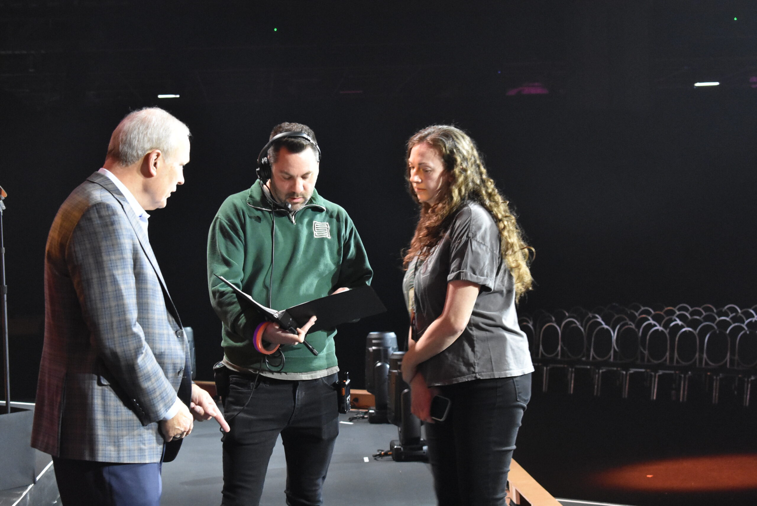 Three people confer onstage, reviewing a list in a binder.