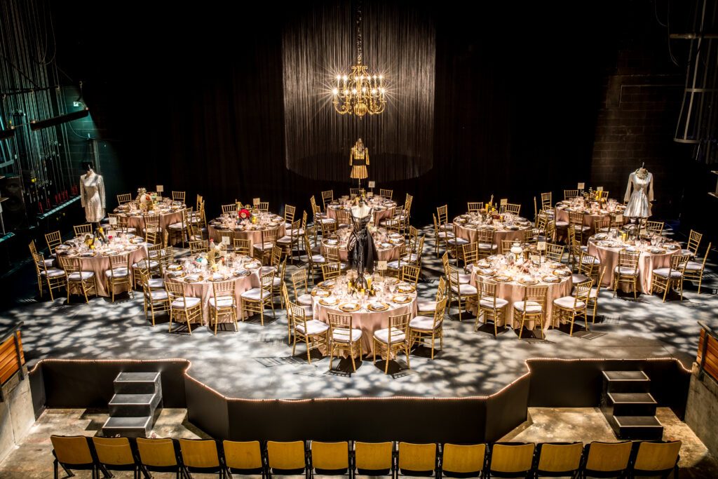 Elegantly-decorated tables stand onstage in a theater, with dresses forming the centerpiece at several tables.