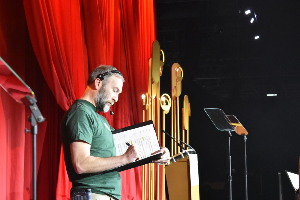 A person stands onstage reviewing a checklist at an event.