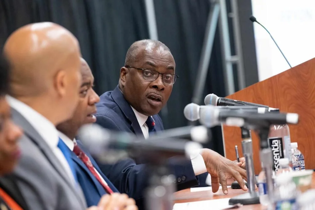 A panelist gestures, looking towards the camera, as he speaks into his mic.