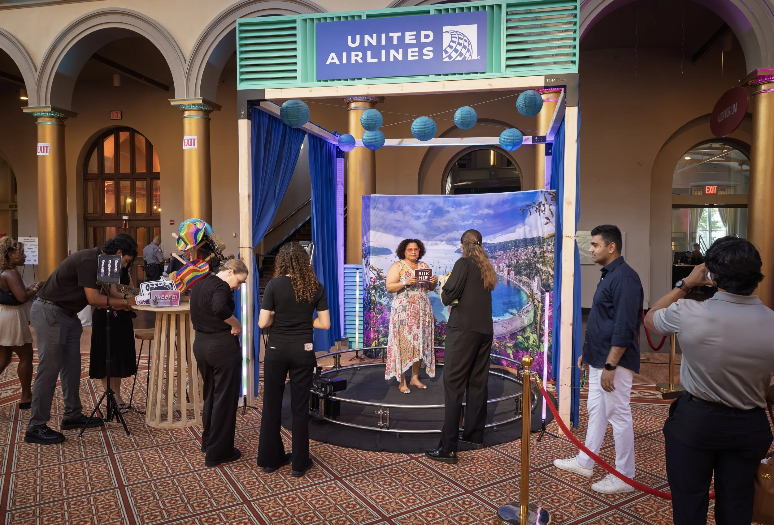 A woman poses for a photo on a small stage with a backdrop as event production staff adjust the setup.