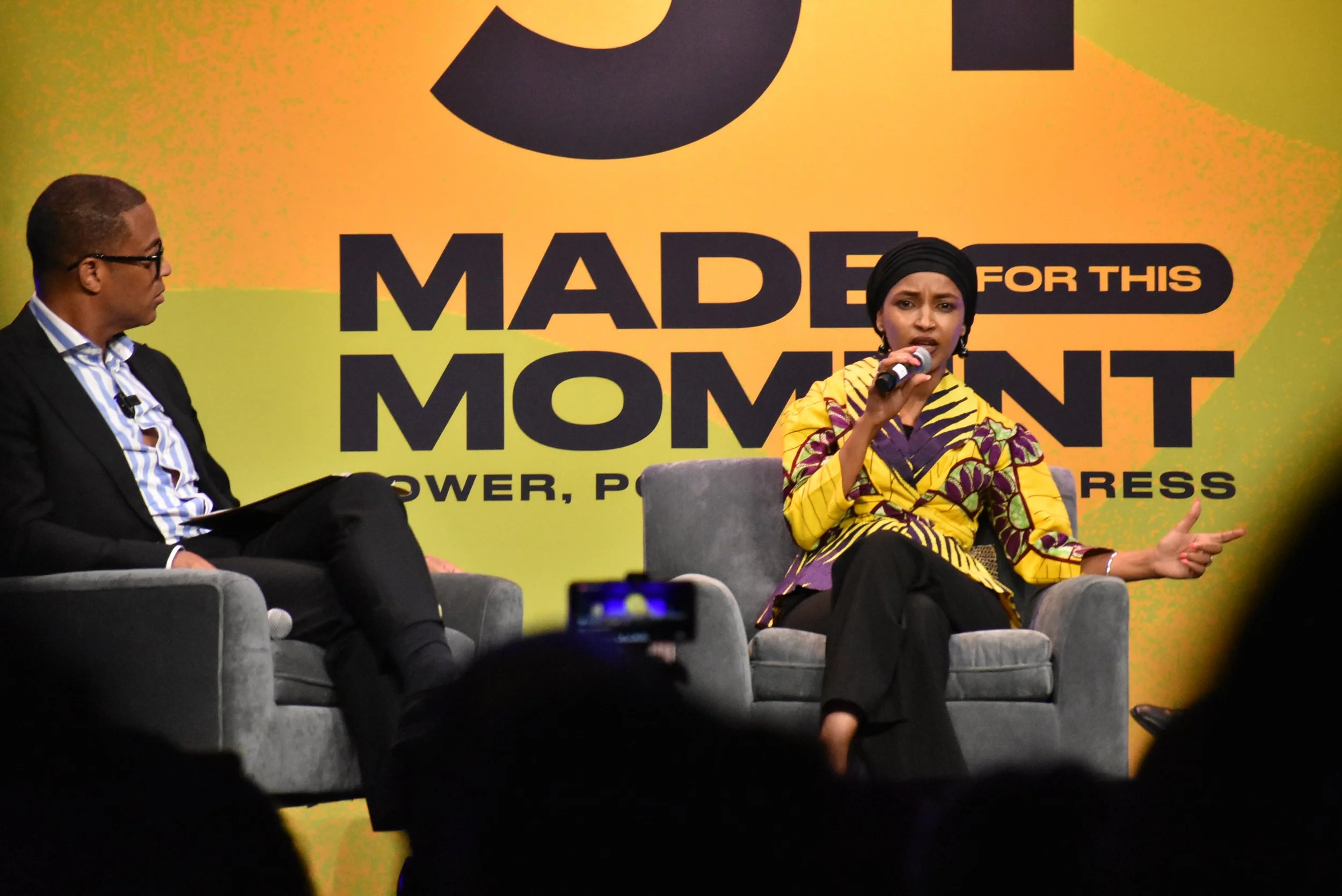 A woman speaks into a mic onstage in front of a screen that reads "Made For This Moment" as a man sitting next to her listens.