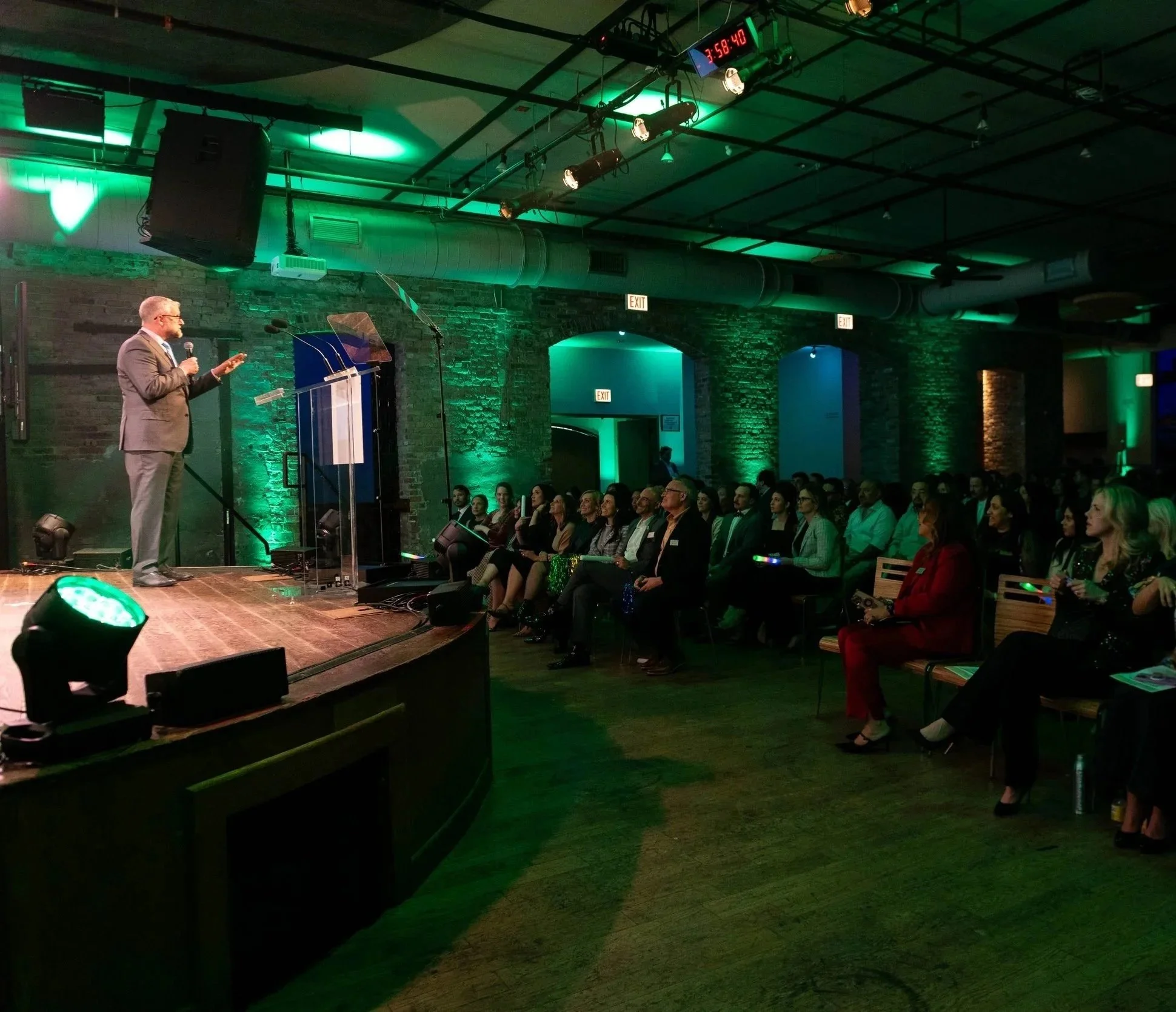 A man in a suit onstage addresses the audience in a hall lit with green lights.