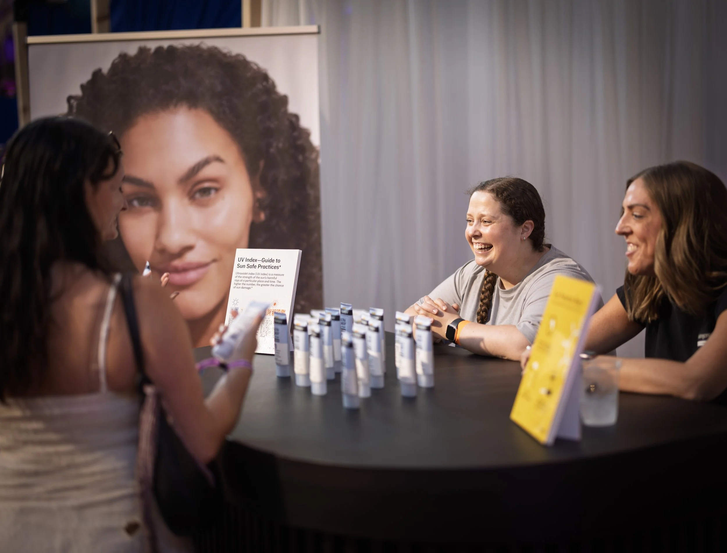 Three people chat with each other over a table of sunscreen samples with a UV index guide on it.